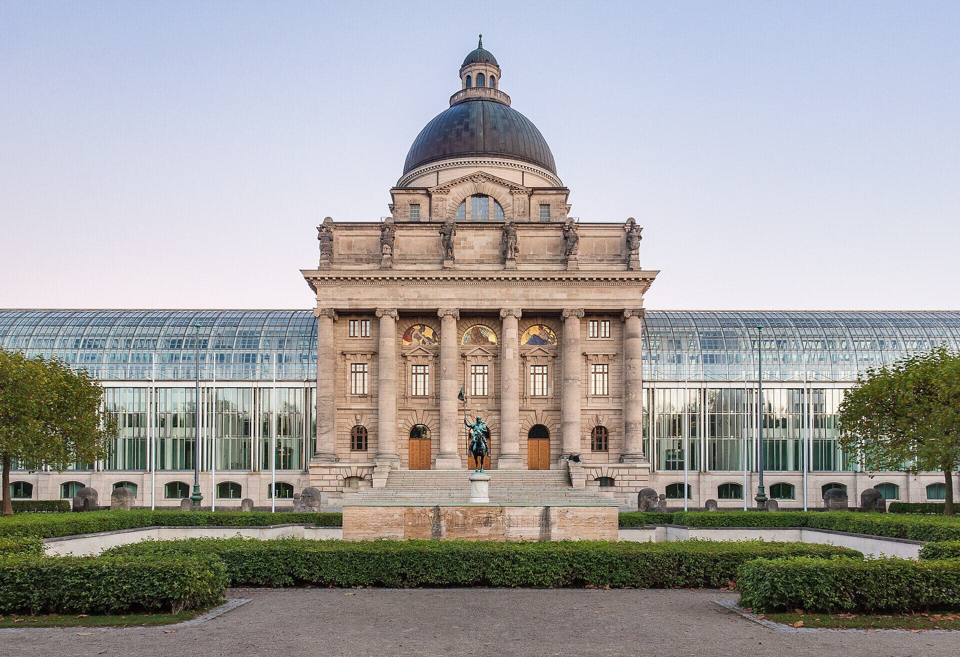 Frontalansicht der Bayerischen Staatskanzlei am Hofgarten in München mit Kuppelbau und Glasflügeln bei klarem Himmel.