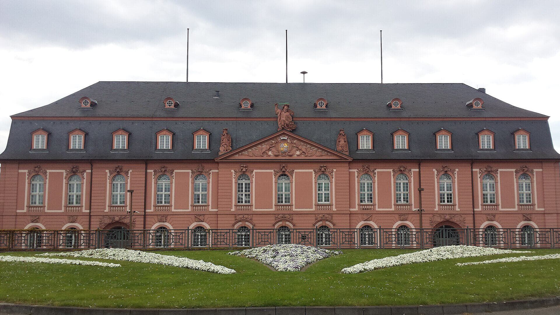 Frontalansicht der Staatskanzlei Rheinland-Pfalz im barocken Neuen Zeughaus in Mainz mit Blumenbeeten im Vordergrund.