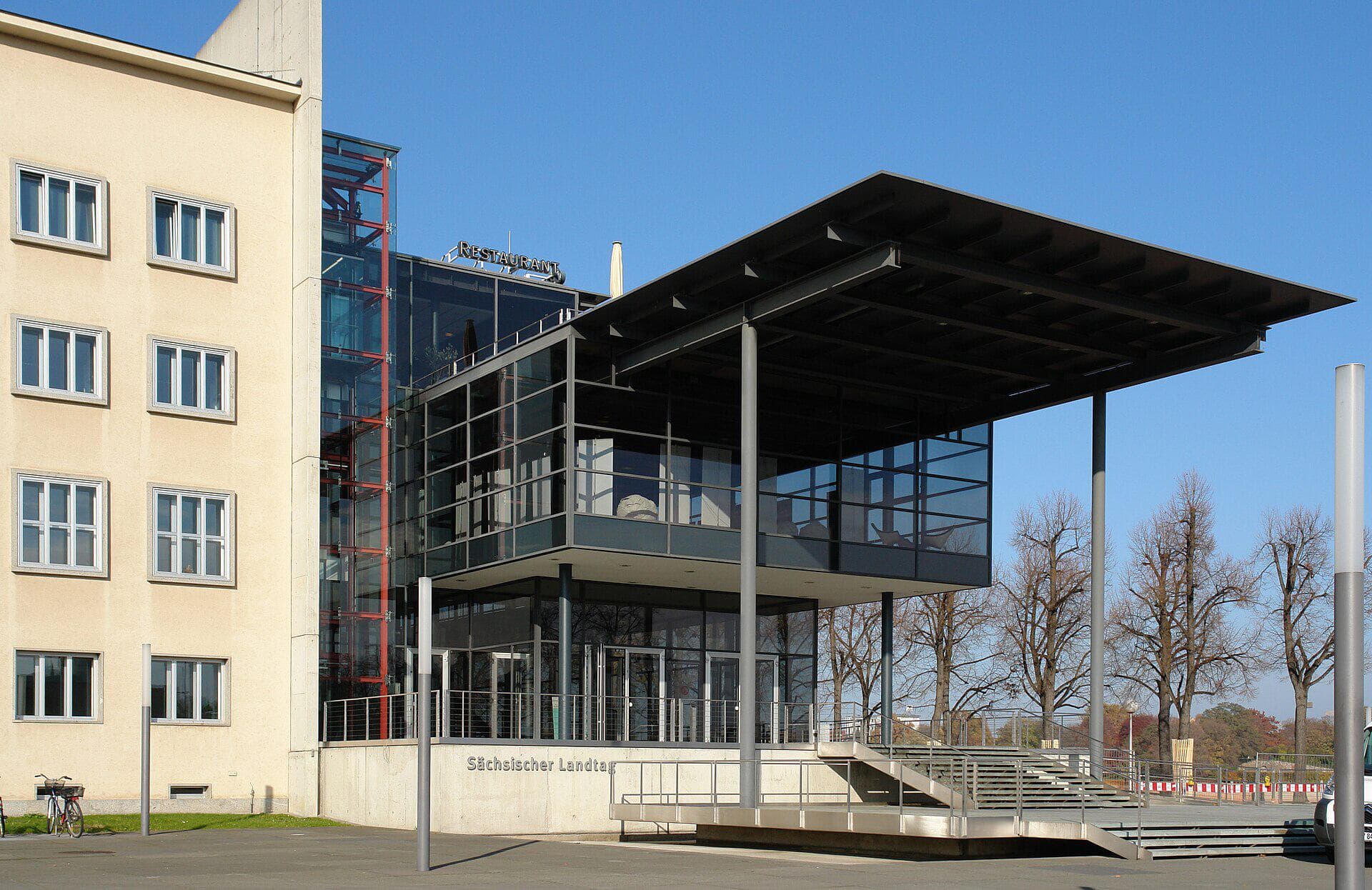Eingangsbereich des Sächsischen Landtags in Dresden mit dem markanten Glasbau und Schriftzug bei blauem Himmel.
